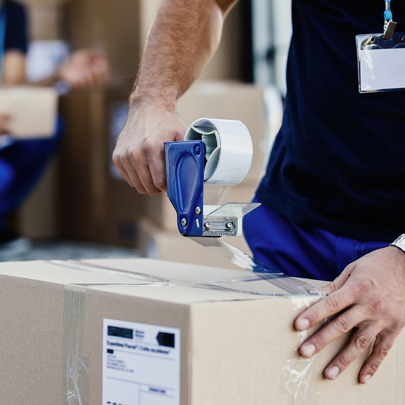 Close-up-of-worker-using-tape-dispenser-while-packing-cardboard-box-for-a-delivery